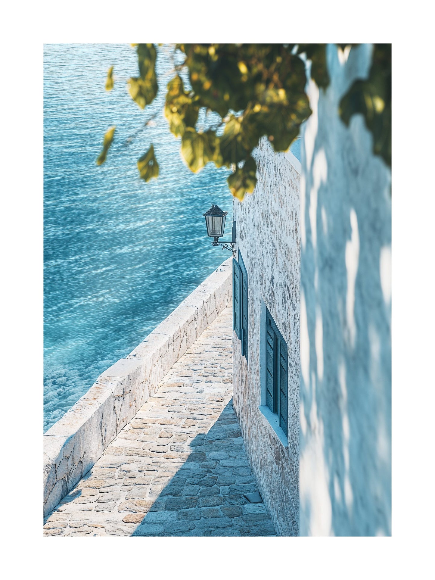 Sunlit narrow walkway by a white stone house overlooking calm ocean water. Ideal for wall art in your home, office, or studio.